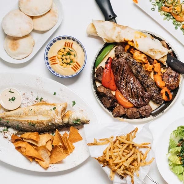 A Lebanese feast with grilled meats, whole fish, hummus, pita, fries, and salad on a white table.